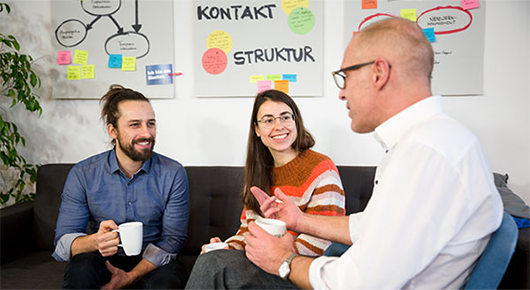 Drei Personen im Gesprch, im Hintergrund Flipchart-Seiten mit Moderationskarten und Notizen