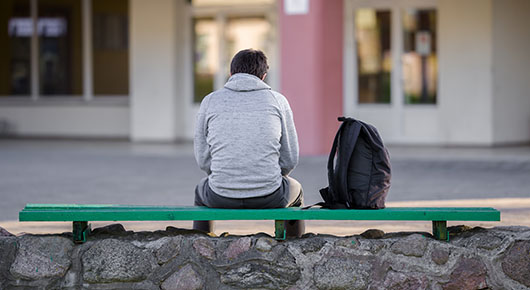 Ein junger Mann sitzt mit dem Rücken zur Kamera auf einer Bank vor einem Gebäude. Neben ihm ein Rucksack.