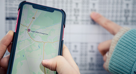 Auf einem Handybildschirm ist eine Straßenkarte zu sehen, im Hintergrund ein Busfahrplan. Auf einem Handybildschirm ist eine Straßenkarte zu sehen, im Hintergrund ein Busfahrplan.