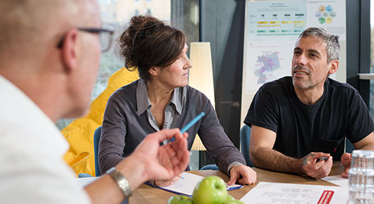 Zwei Männer und eine Frau sitzen gemeinsam an einem Tisch, auf dem Arbeitsunterlagen liegen. Zwei Männer und eine Frau sitzen gemeinsam an einem Tisch, auf dem Arbeitsunterlagen liegen.