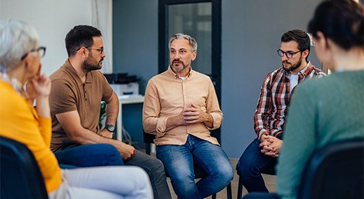 Eine Gruppe von Personen sitzt in einem Stuhlkreis zusammen. Ein Mann spricht, die anderen hören interessiert zu. Eine Gruppe von Personen sitzt in einem Stuhlkreis zusammen. Ein Mann spricht, die anderen hören interessiert zu.