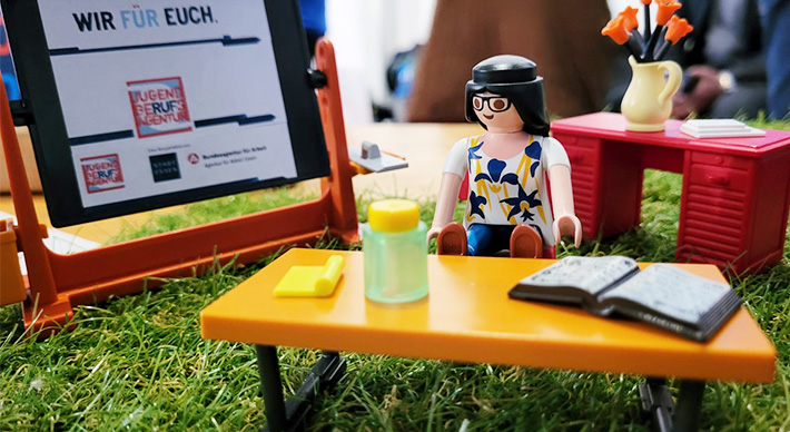 Lego-Modell mit Figur an Schreibtisch sowie Wandtafel mit Logo der JBA Essen und Text "Wir für euch" Lego-Modell mit Figur an Schreibtisch sowie Wandtafel mit Logo der JBA Essen und Text "Wir für euch"