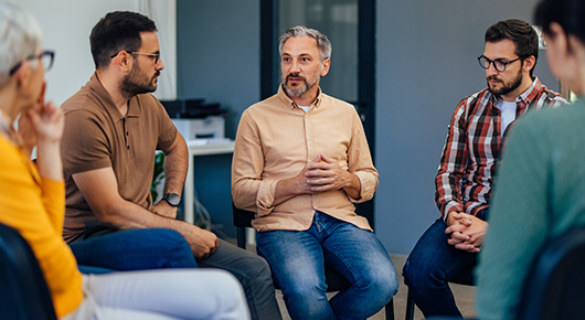 Eine Gruppe von Personen sitzt in einem Stuhlkreis zusammen. Ein Mann spricht, die anderen hören interessiert zu. Eine Gruppe von Personen sitzt in einem Stuhlkreis zusammen. Ein Mann spricht, die anderen hören interessiert zu.