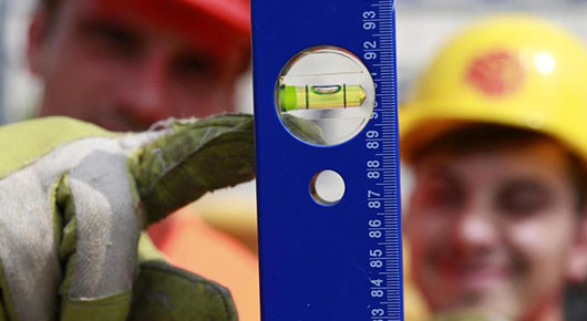 Im Vordergrund eine blaue Wasserwaage, auf die eine Hand im Arbeitshandschuh zeigt. Im Hintergrund ein Mann mit Schutzhelm.