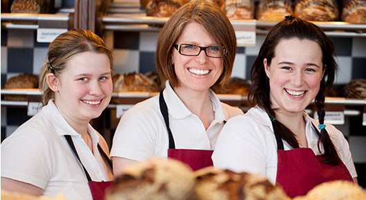 Drei Frauen mit weißen Blusen und roten Schürzen stehen hinter einem Tresen. Im Hintergrund Regale mit Broten.